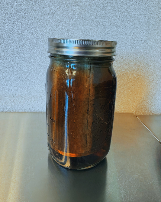 A glass mason jar with dark golden brown tea and a steel mesh infuser inside sits on a stainless steel prep table.